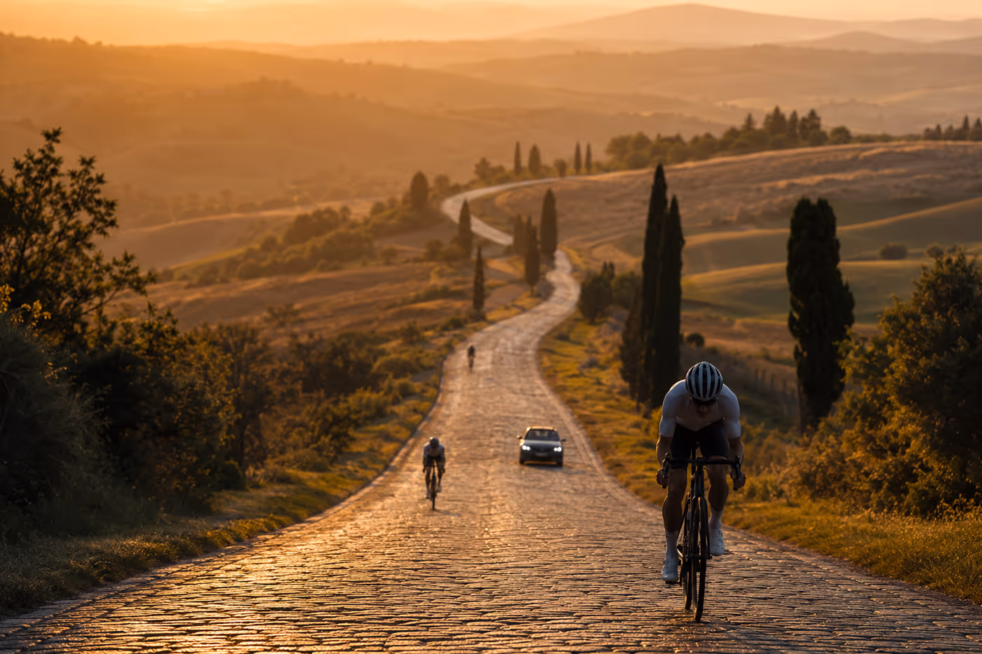 A cobbled road winding between hills at sunset, three riders hundreds of metres apart and a support car in one of the gaps — the race has stopped being a peloton.